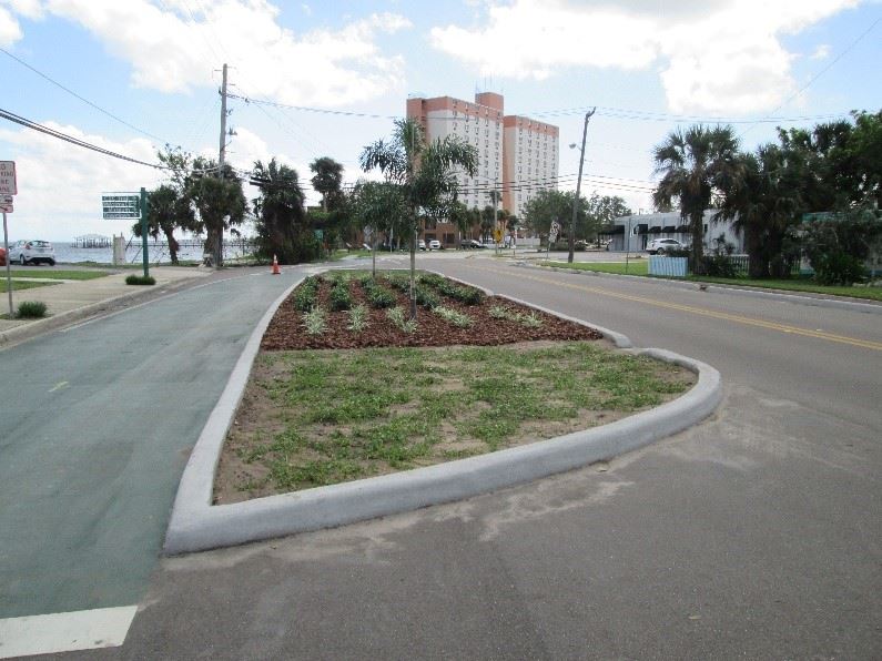 Photo of a curbed island of grass and mulch in the middle of a paved roadway.