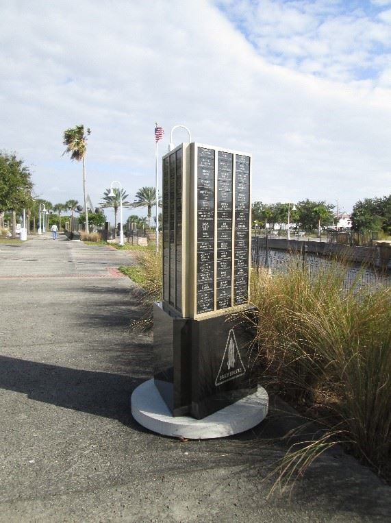 Photo of a triangular-shaped monument, with names inscribed on all sides, next to a body of water.